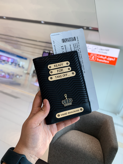 A hand holding a black personalized passport cover with text on it, displayed in an airport setting.