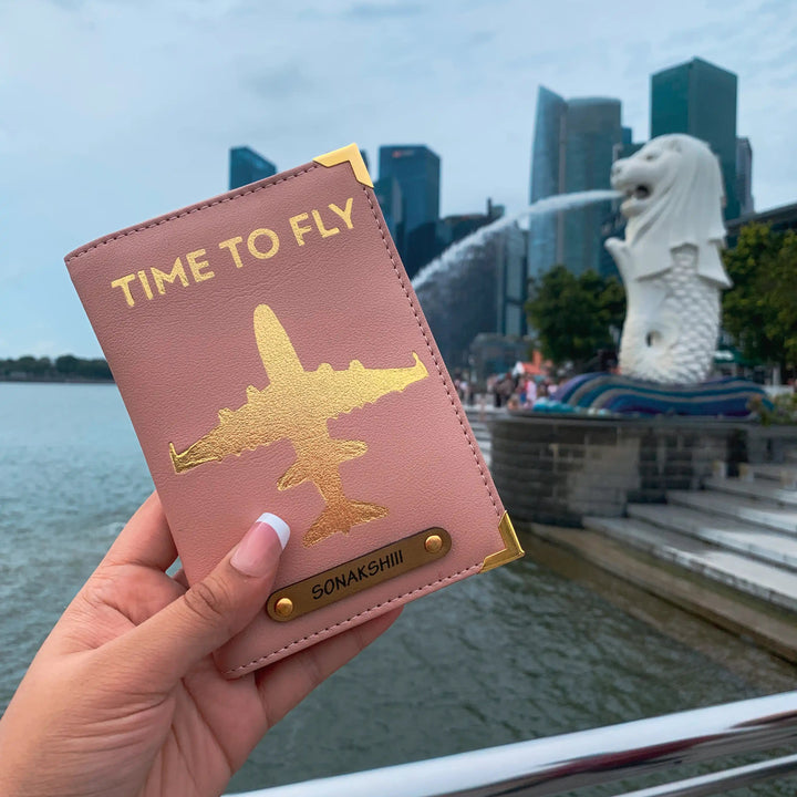 A hand holding a pink passport cover with gold aviation-inspired design and text &