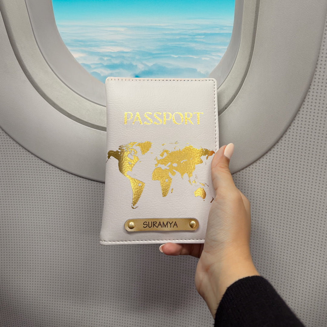 Hand holding a passport cover with a world map design in front of an airplane window. Brand TPC Gifts