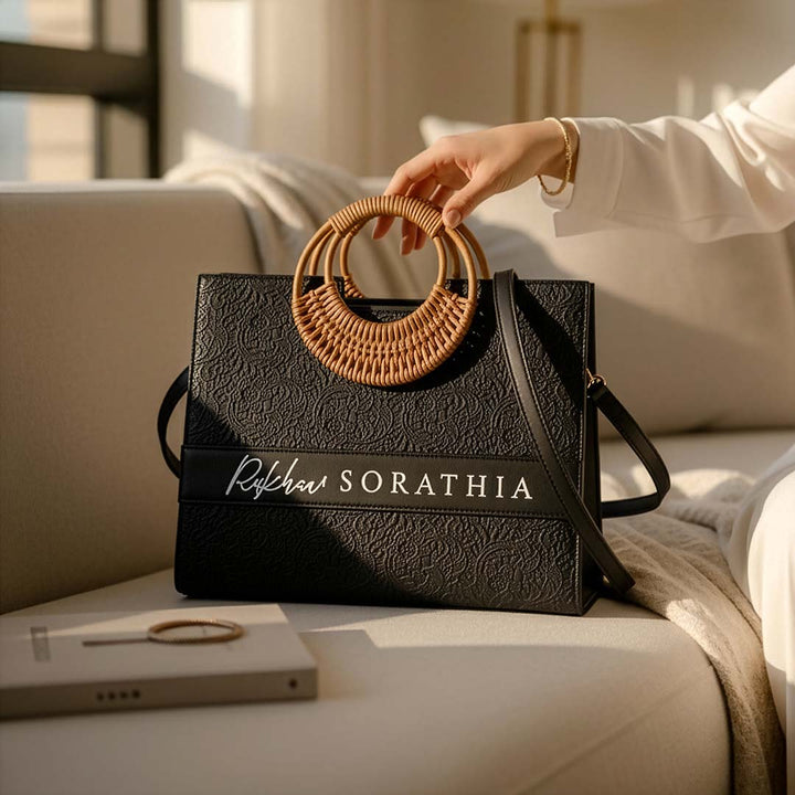 Black handbag with woven handle held by a hand on a light-colored couch.