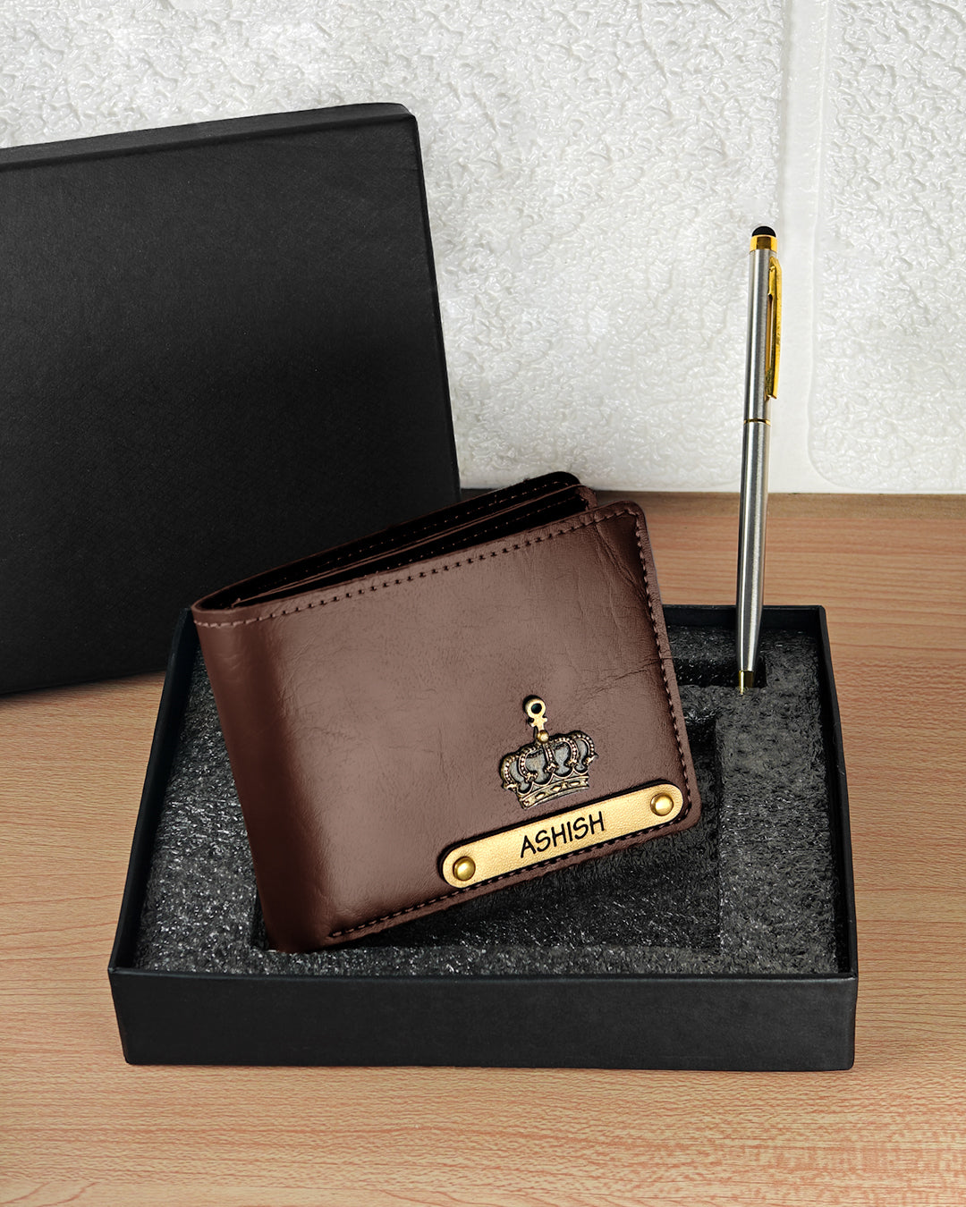 Brown wallet with 'ASHISH' branding on a black stand with a pen and black box in the background - TPC Gifts
