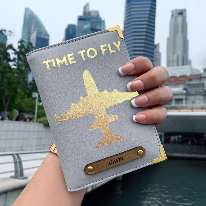 A hand holding a grey passport cover with gold aviation-inspired design and text &