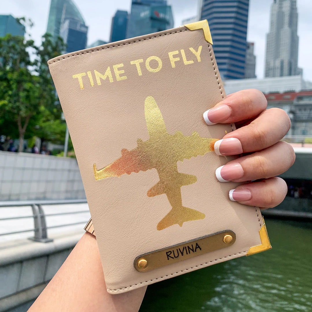 A hand holding a beige passport cover with gold aviation-inspired design and text &