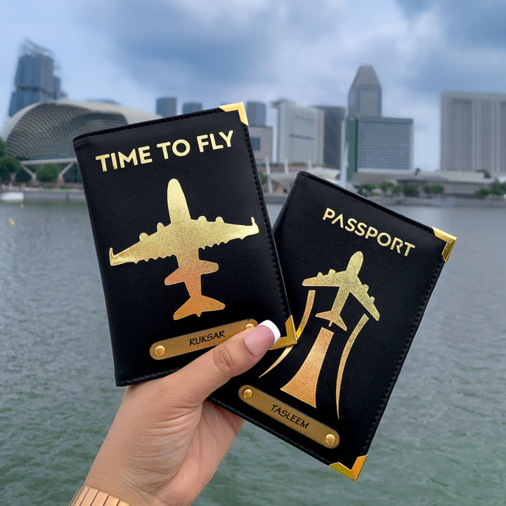 Two passport covers held in hand, one black and one beige, with a gold aviation-inspired design and text &