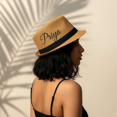 Woman wearing a straw hat with 'Priya' embroidered on it, standing against a beige wall with shadow patterns.