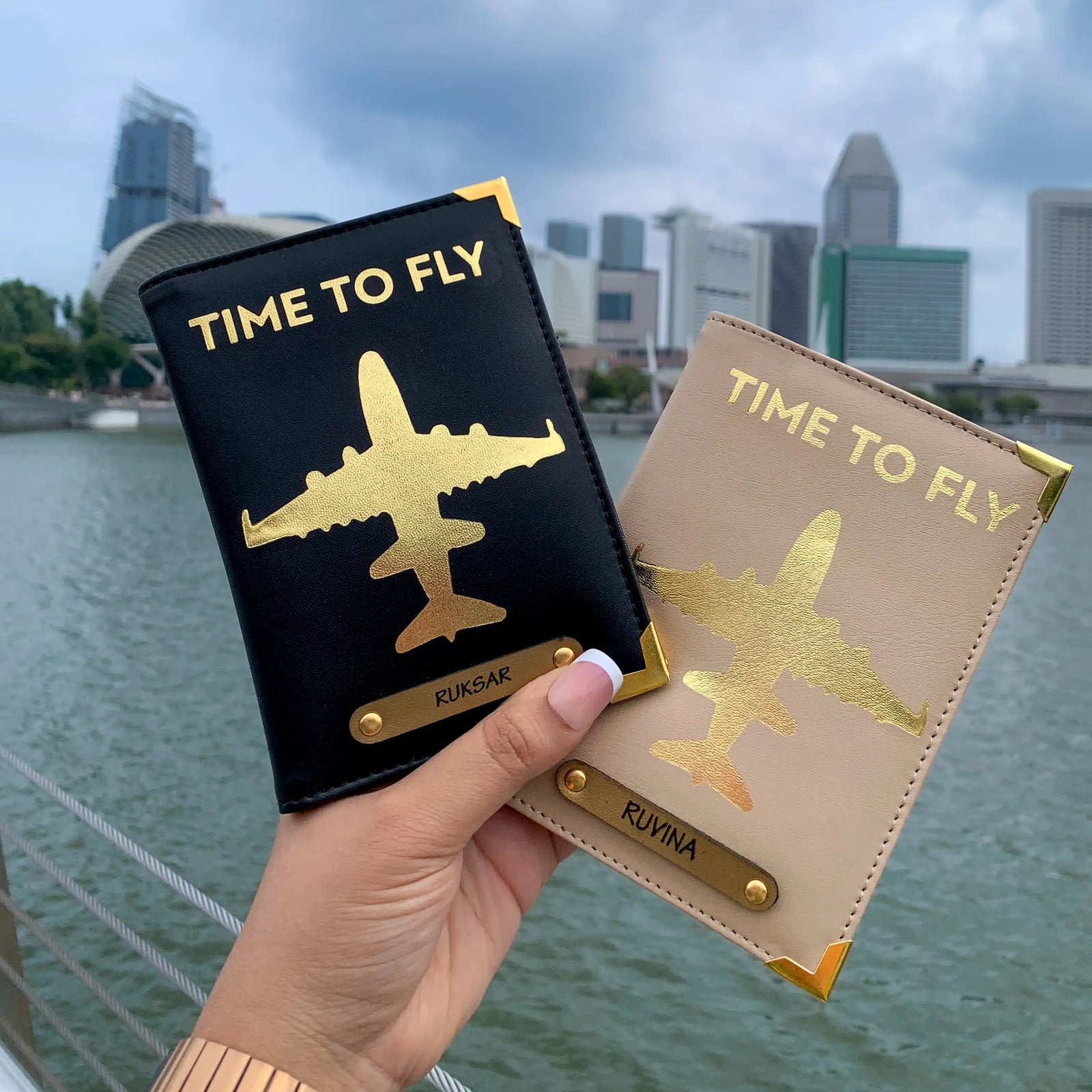 Two passport covers held in hand, one black and one beige, with a gold aviation-inspired design and text 'TIME TO FLY' on the black cover, displayed against a scenic background.