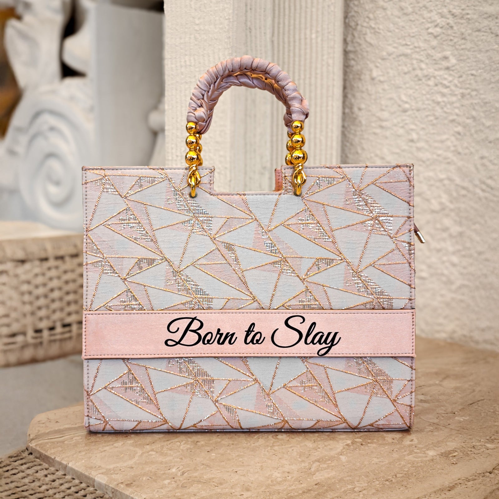A tote bag with a geometric pattern in pink tones and a gold-plated handle, placed on a marble surface with the text 'Born to Slay' on the front.