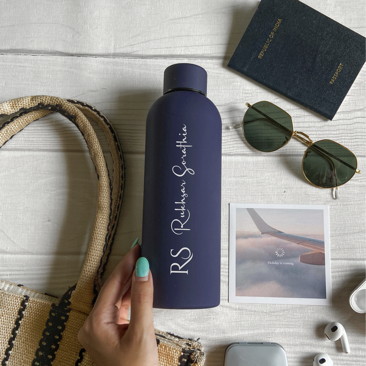 A personalized blue stainless steel water bottle with custom engraving, displayed alongside a passport, sunglasses, and earphones on a white surface.