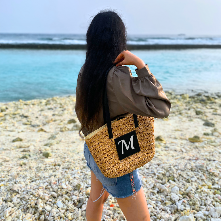 Model carrying the Personalised Brown Cane Beach Bag - Luxe Edition on the Maldives beach surrounded by pebbles