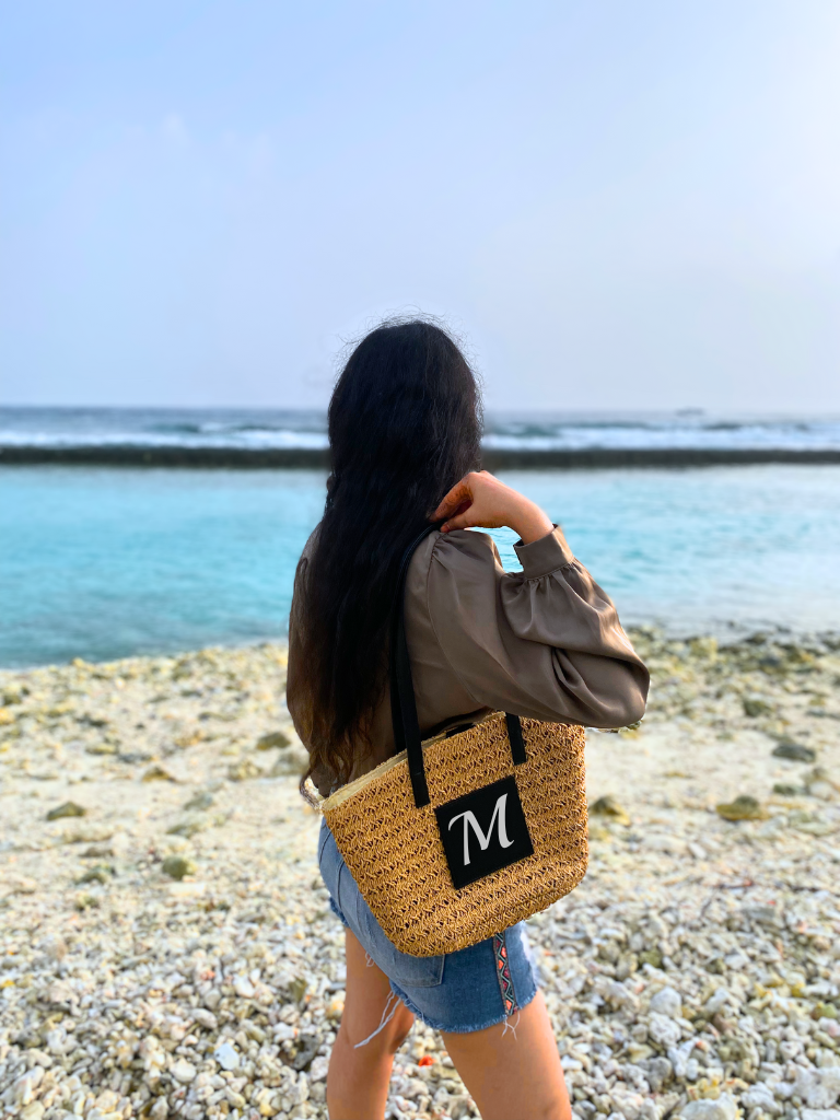 Model carrying the Personalised Brown Cane Beach Bag - Luxe Edition on the Maldives beach surrounded by pebbles