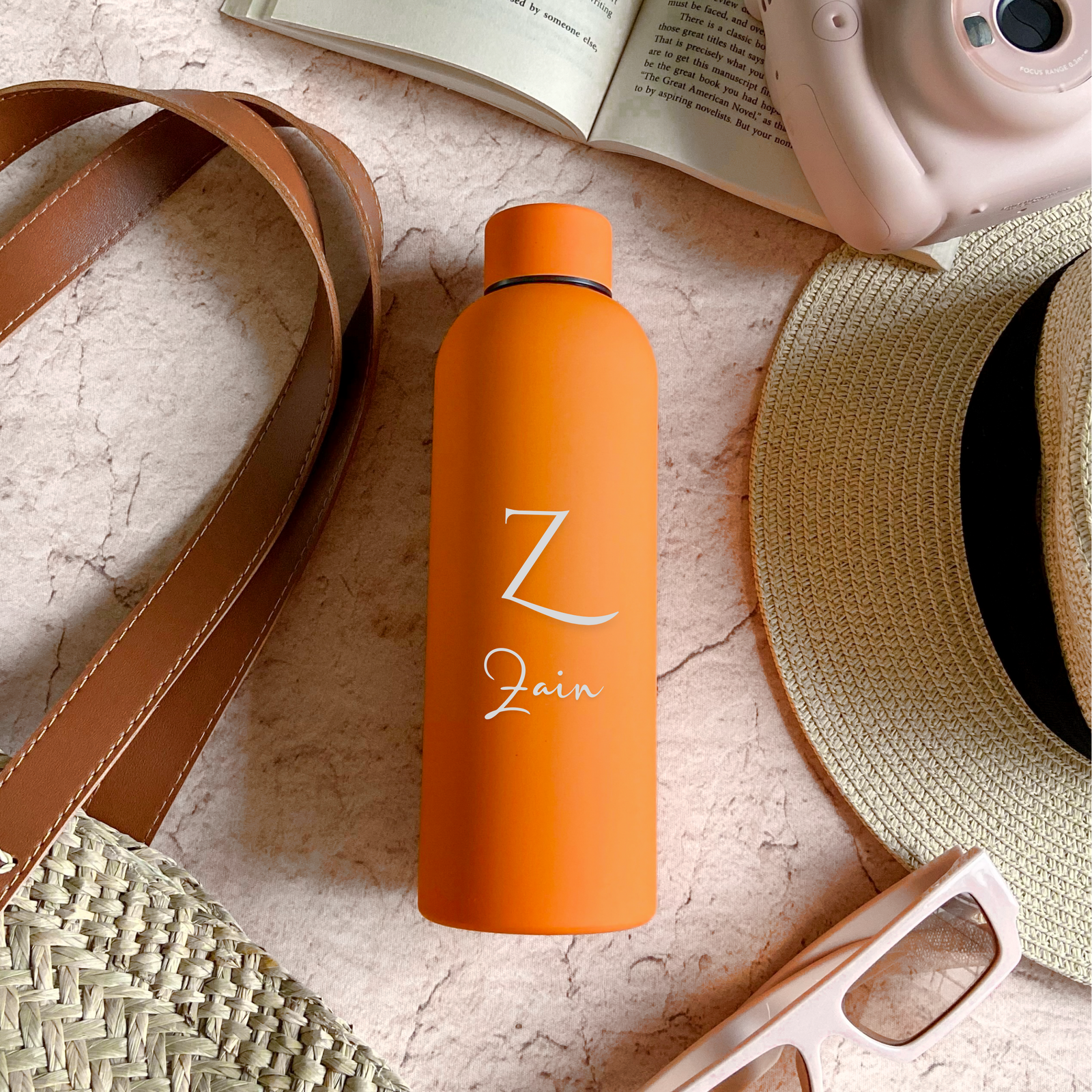 An orange stainless steel water bottle with custom engraving, placed on a textured surface alongside a brown hat, sunglasses, and a camera.