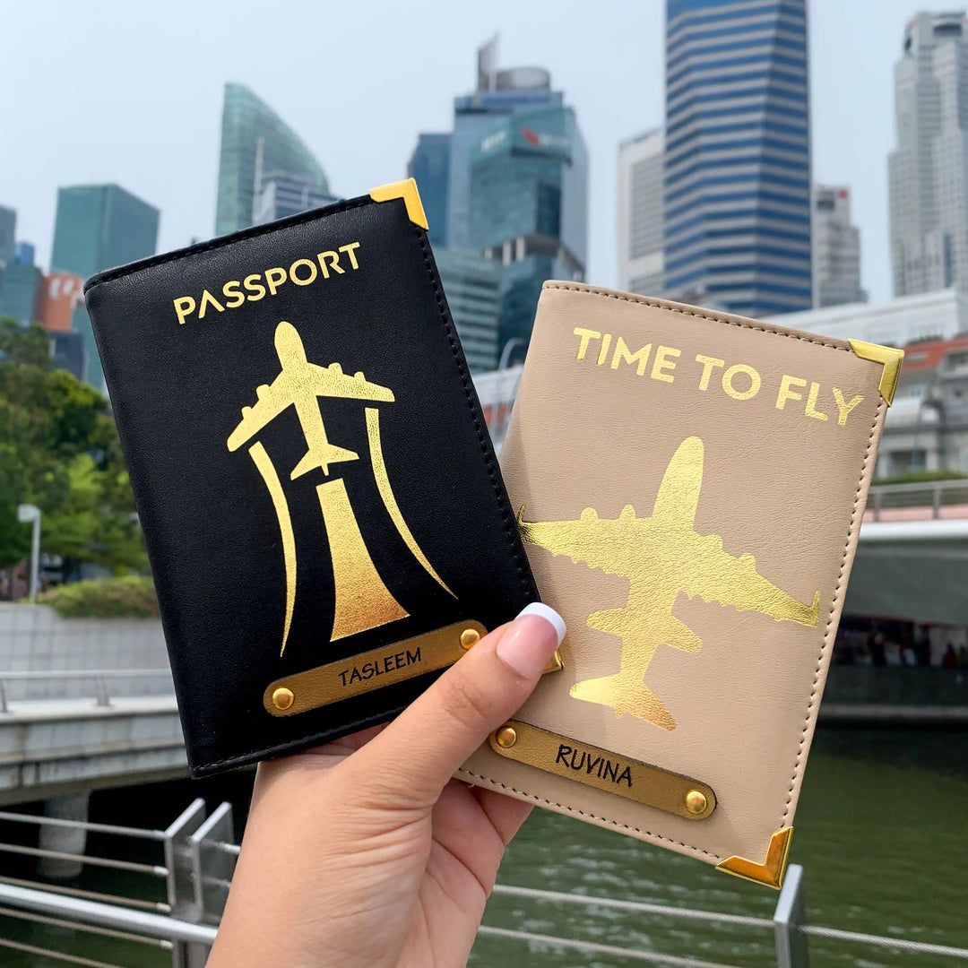 Two passport covers held in hand, one black and one beige, with a gold aviation-inspired design and text &