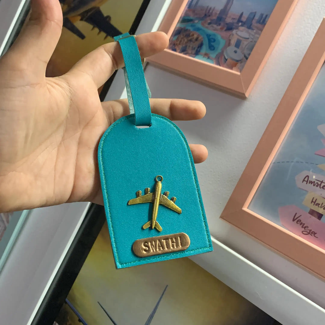 A turquoise blue luggage tag with a gold airplane charm and a personalized nameplate reading &