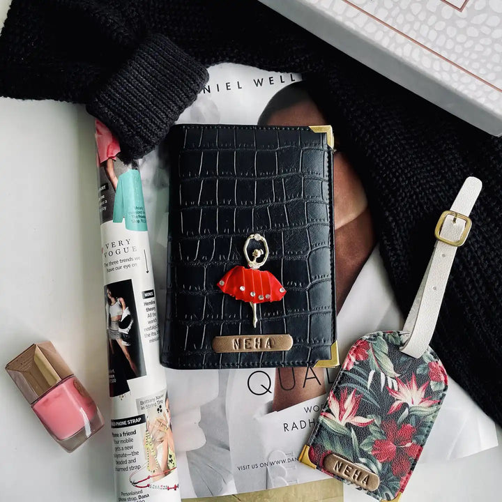Black Croco Passport Cover &amp; Tropical Luggage Tag.A styled flat-lay featuring a black crocodile-embossed passport cover with a red dollvcharm and a matching floral luggage tag, both branded NEHA, alongside cosmetics and a black knit sweater on a white surface-TPC Gifts