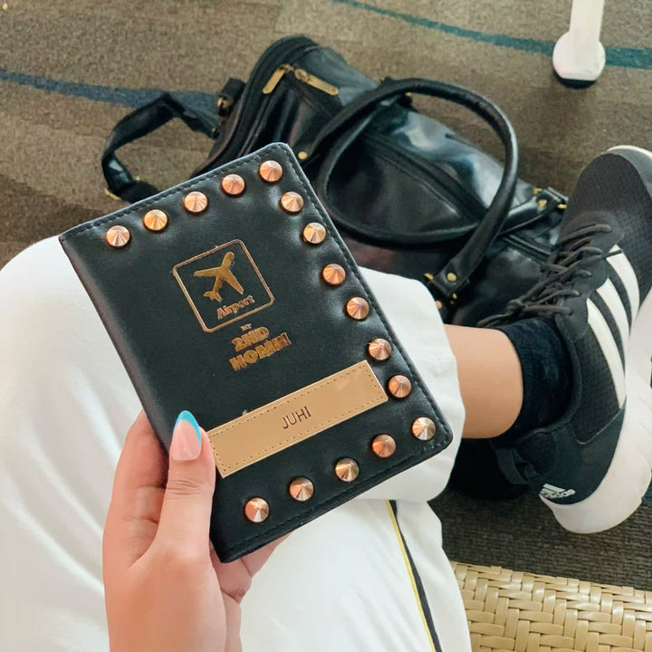 A person holds a black faux-leather passport cover with rose gold studs and a gold-toned airplane logo. The scene is a casual indoor setting with a black handbag and a black-and-white sneaker visible in the background-TPC Gifts