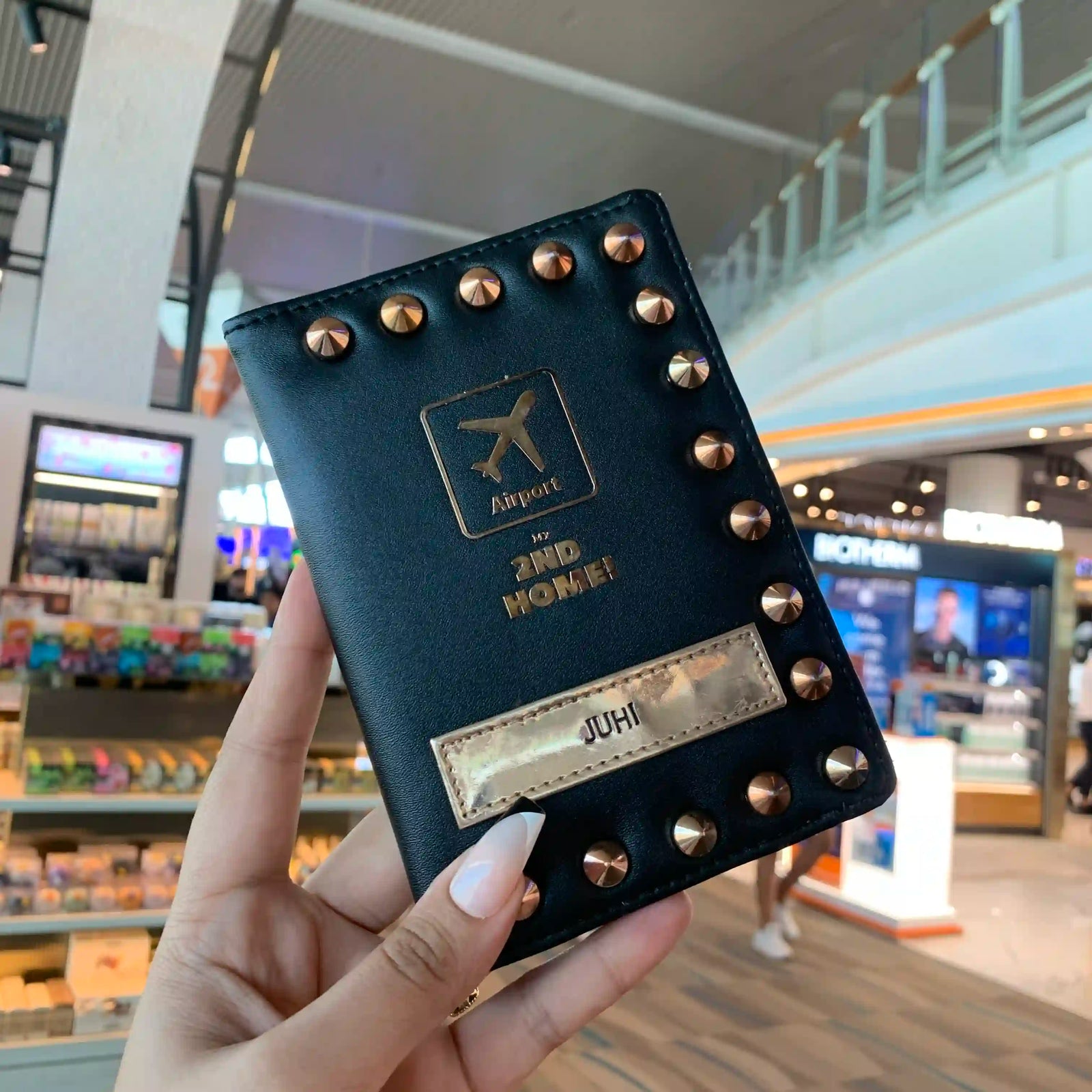 A hand holds a black faux-leather passport cover in an airport setting. The cover features gold-tone metal studs around the border, a gold airplane icon with the text 'Airport' and '2ND HOME!' on the front, and a gold nameplate that reads 'JUHJ'.