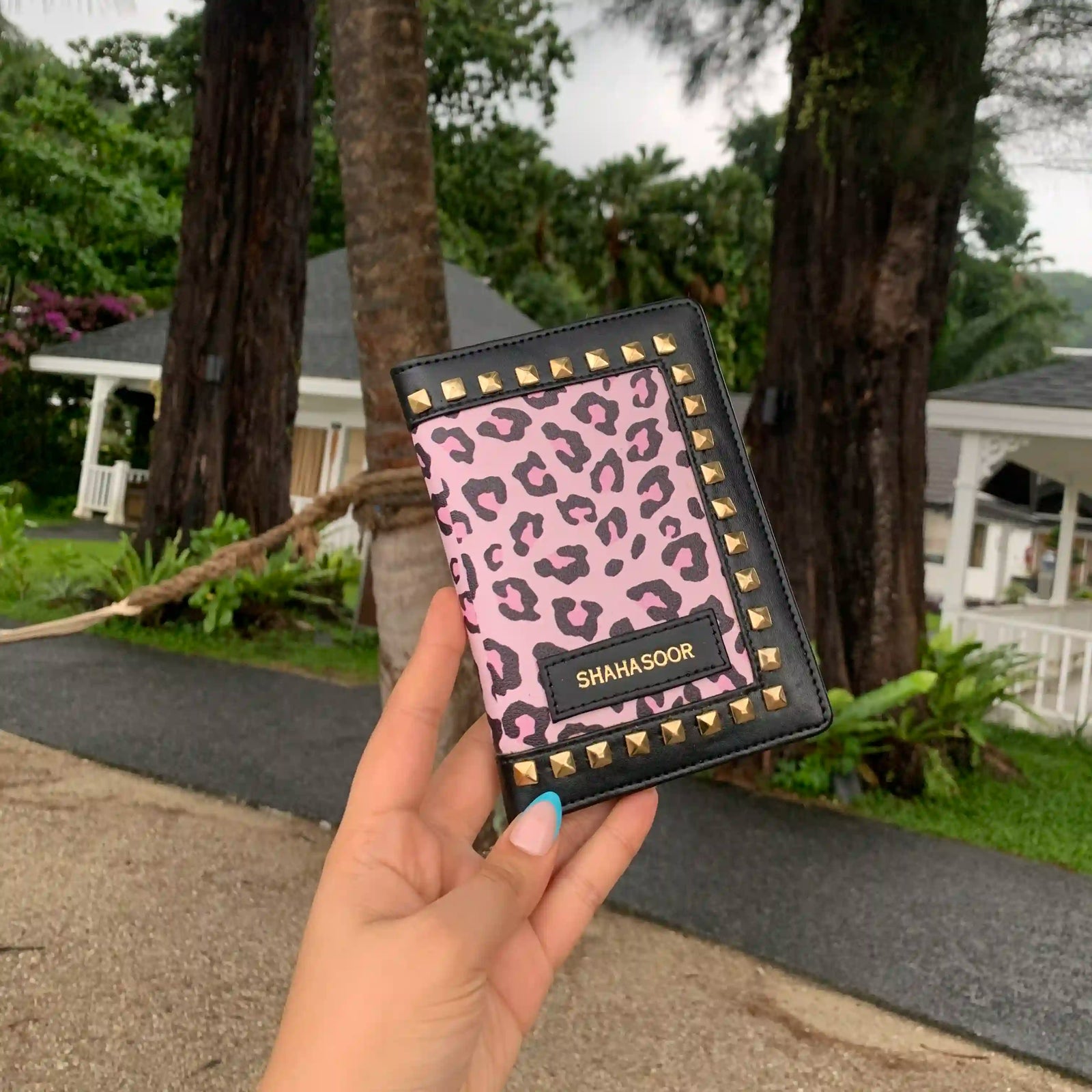 A hand holding a black and pink animal print passport cover with stud details, displayed in front of a garden backdrop -TPC Gifts
