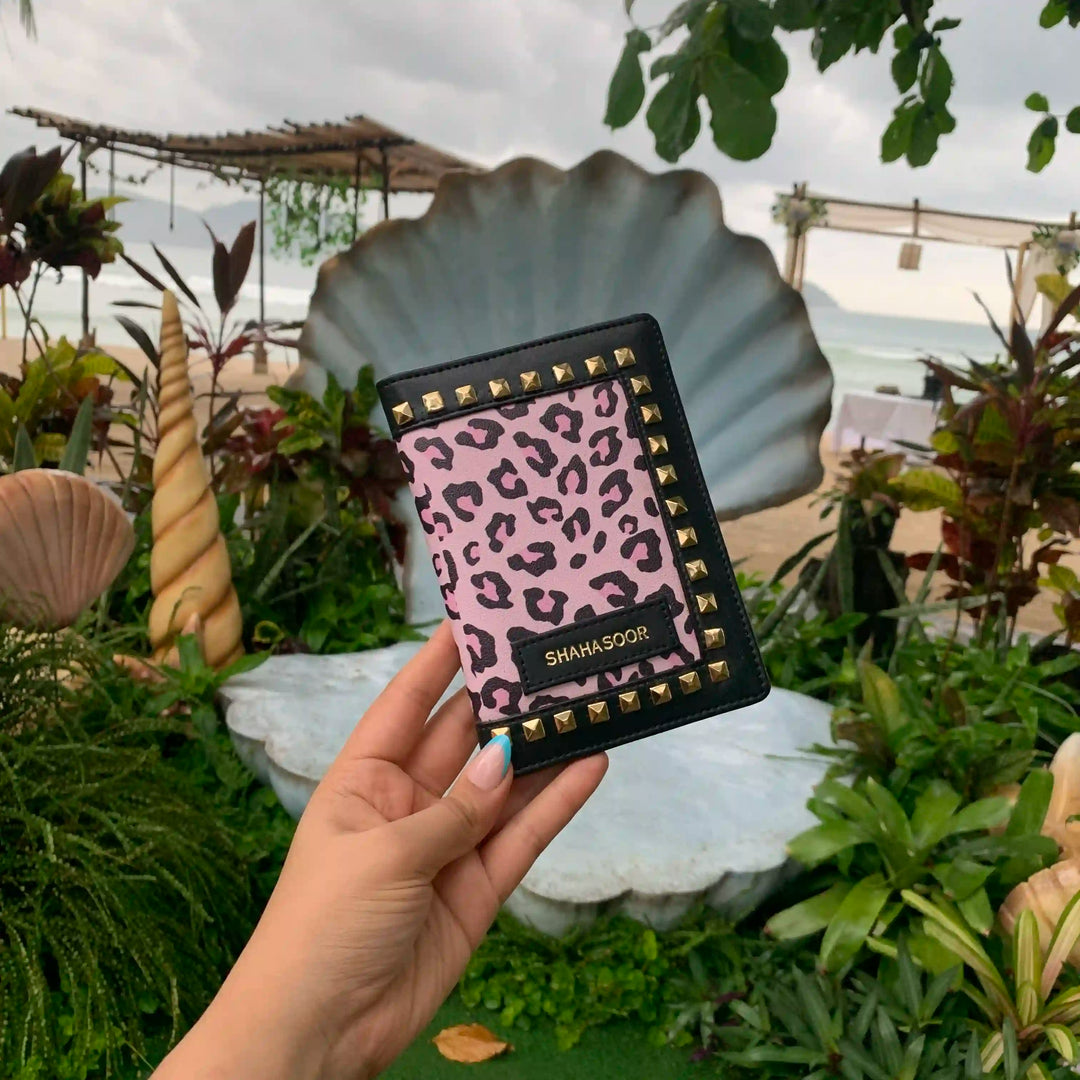 A hand holding a black and pink animal print passport cover with stud details, displayed in front of a garden backdrop -TPC Gifts