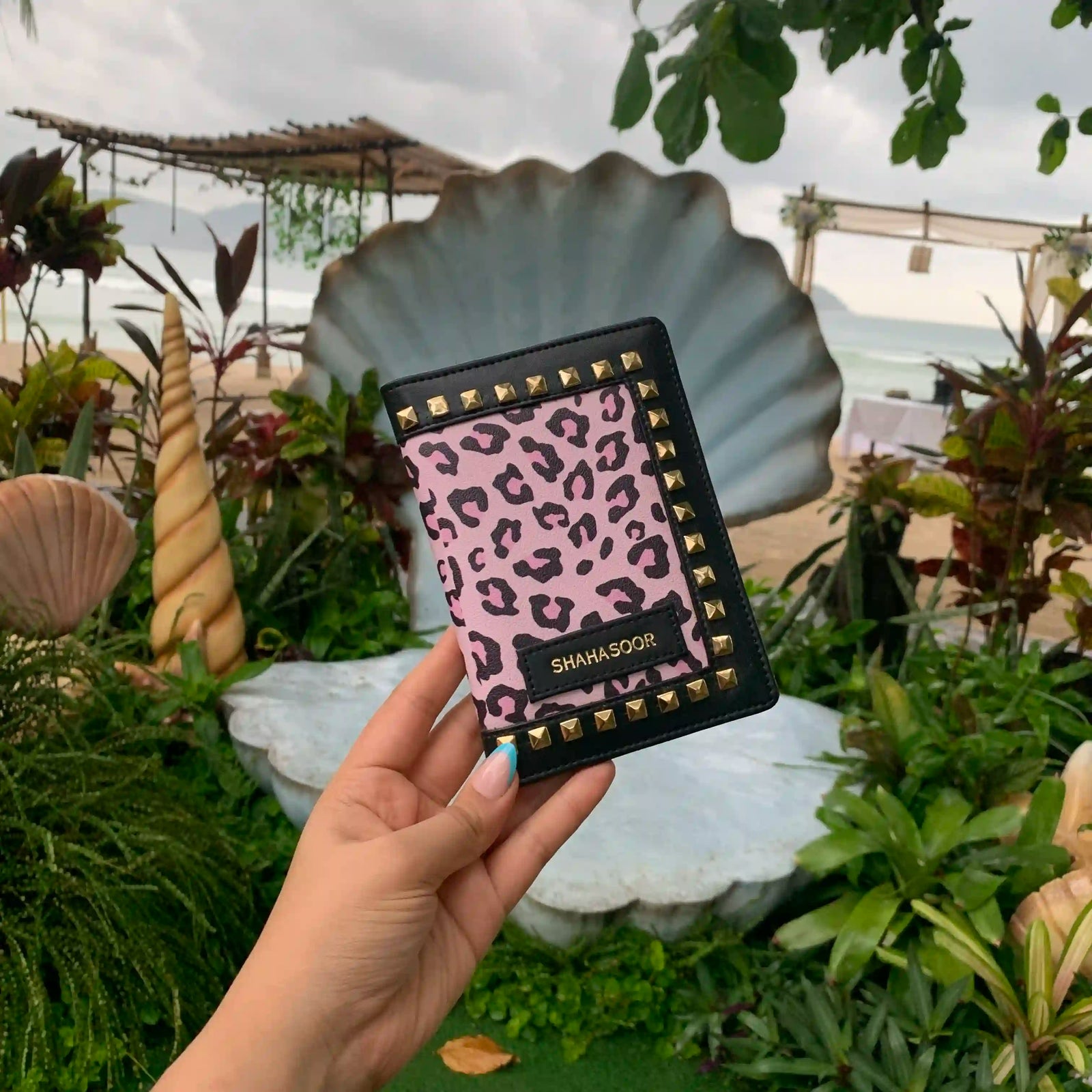 A hand holding a black and pink animal print passport cover with stud details, displayed in front of a garden backdrop -TPC Gifts