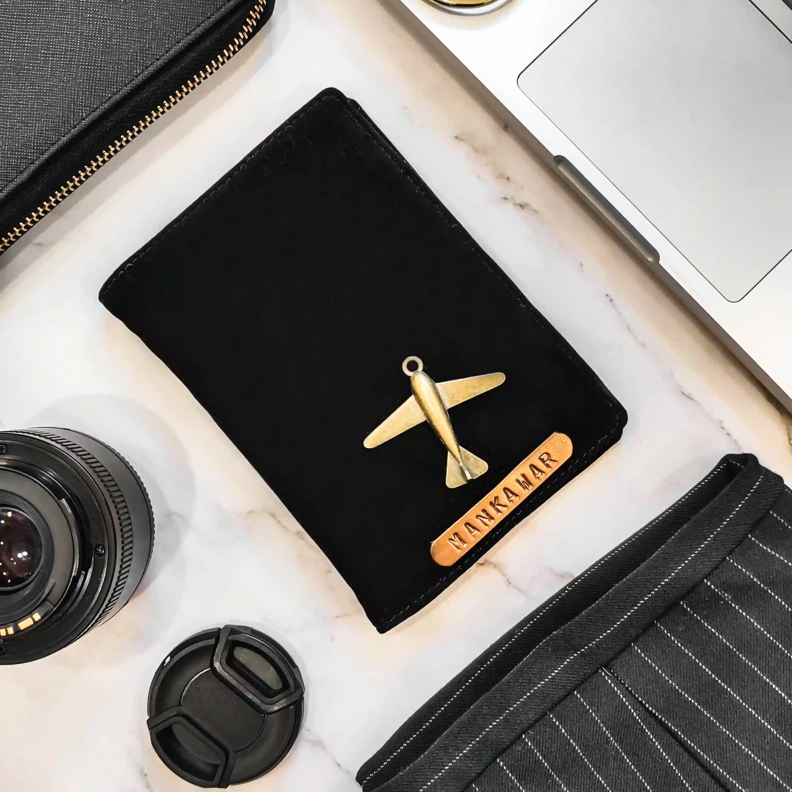 A black velvet passport cover with a gold airplane charm and a personalized wooden nameplate reading 'HANKAMAR', styled in a flat-lay on a white marble surface with a laptop, camera lens, and other accessories - TPC Gifts