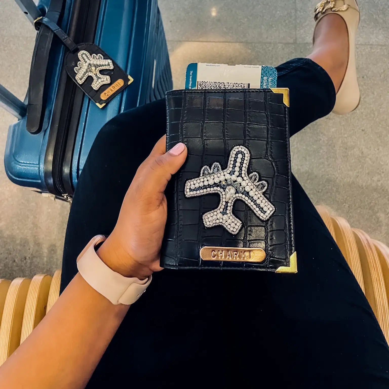 A person sitting with luggage beside them, holding a black passport cover with a crocodile pattern and a name tag. There is also a small white charm with a plane design on it -TPC Gifts