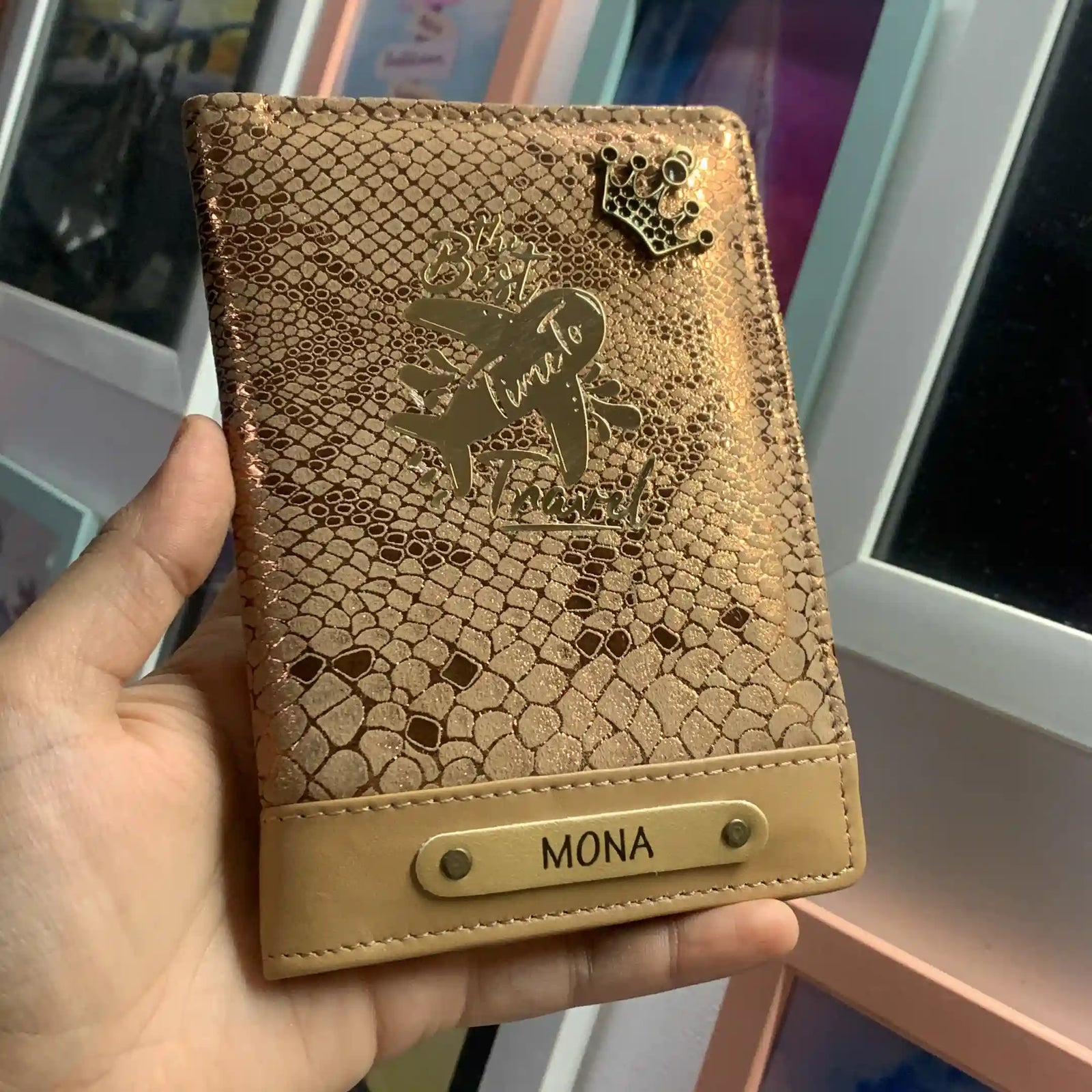 A hand holding a personalized snakeskin-patterned passport cover in a warm tan color. The cover features an embossed design with a crown, the phrase 'THE BEST TIME TO TRAVEL', and the name 'MONA' on a tan leather-like band at the bottom.