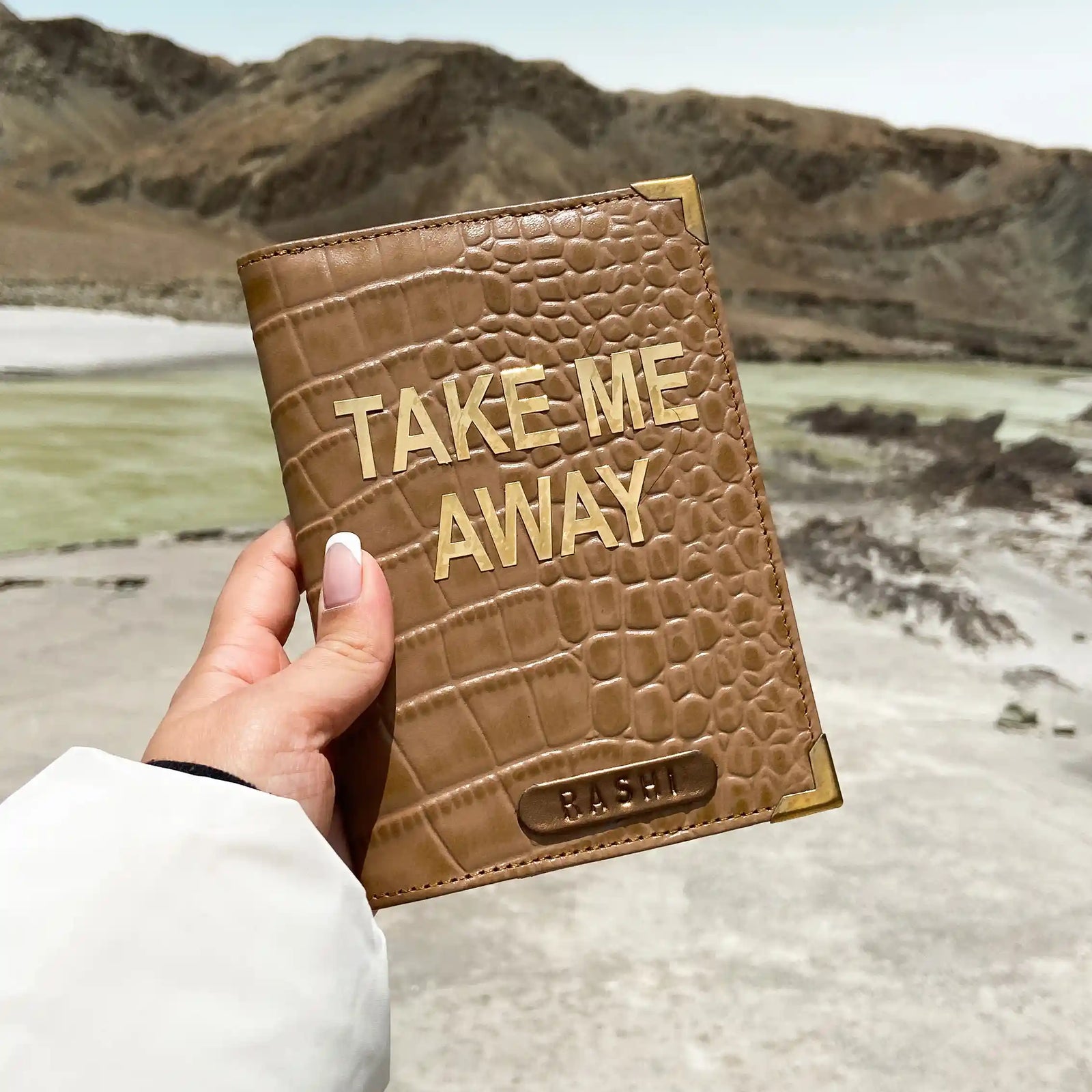 A passport cover with a crocodile pattern in a buff cream color, held up in a landscape setting. - TPC Gifts