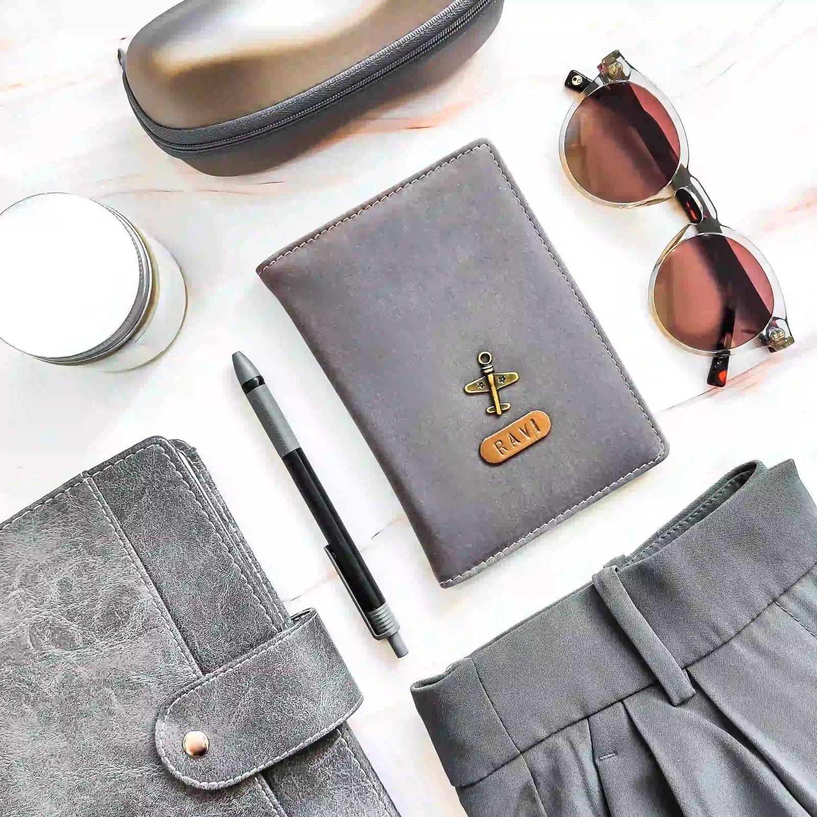 A styled flat-lay featuring a gray VELVET passport holder surrounded by a gray zippered case, sunglasses, a pen, a candle, and gray fabric on a white marble background