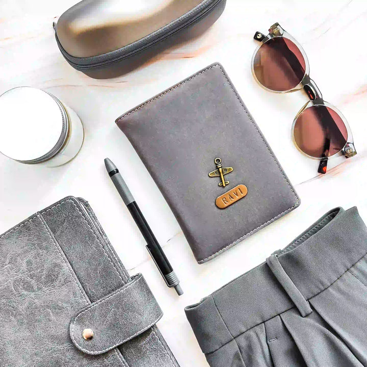 A styled flat-lay featuring a gray VELVET passport holder surrounded by a gray zippered case, sunglasses, a pen, a candle, and gray fabric on a white marble background
