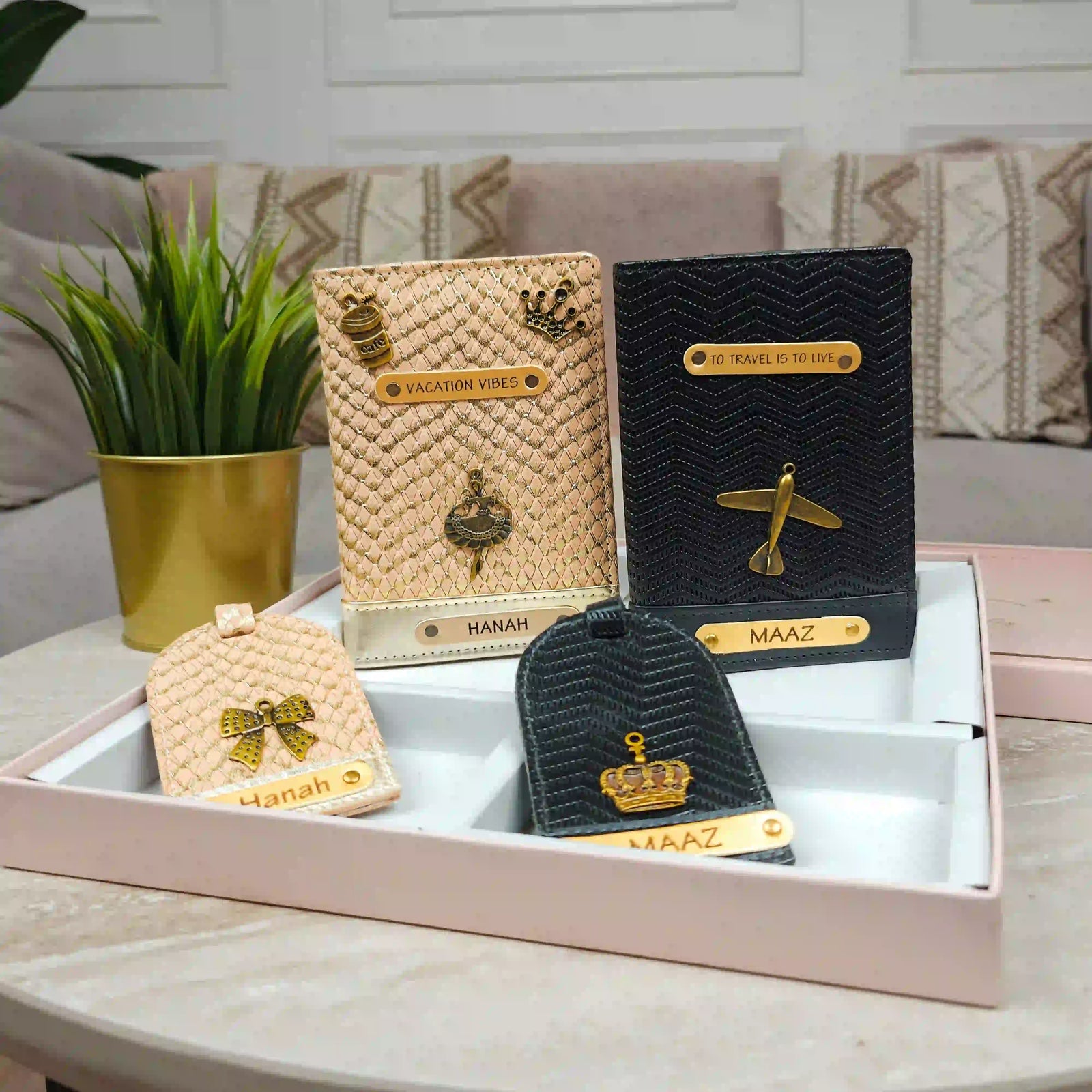 A set of personalized luggage tags and passport covers displayed in a pink presentation box on a marble table, with a potted plant and sofa in the background.