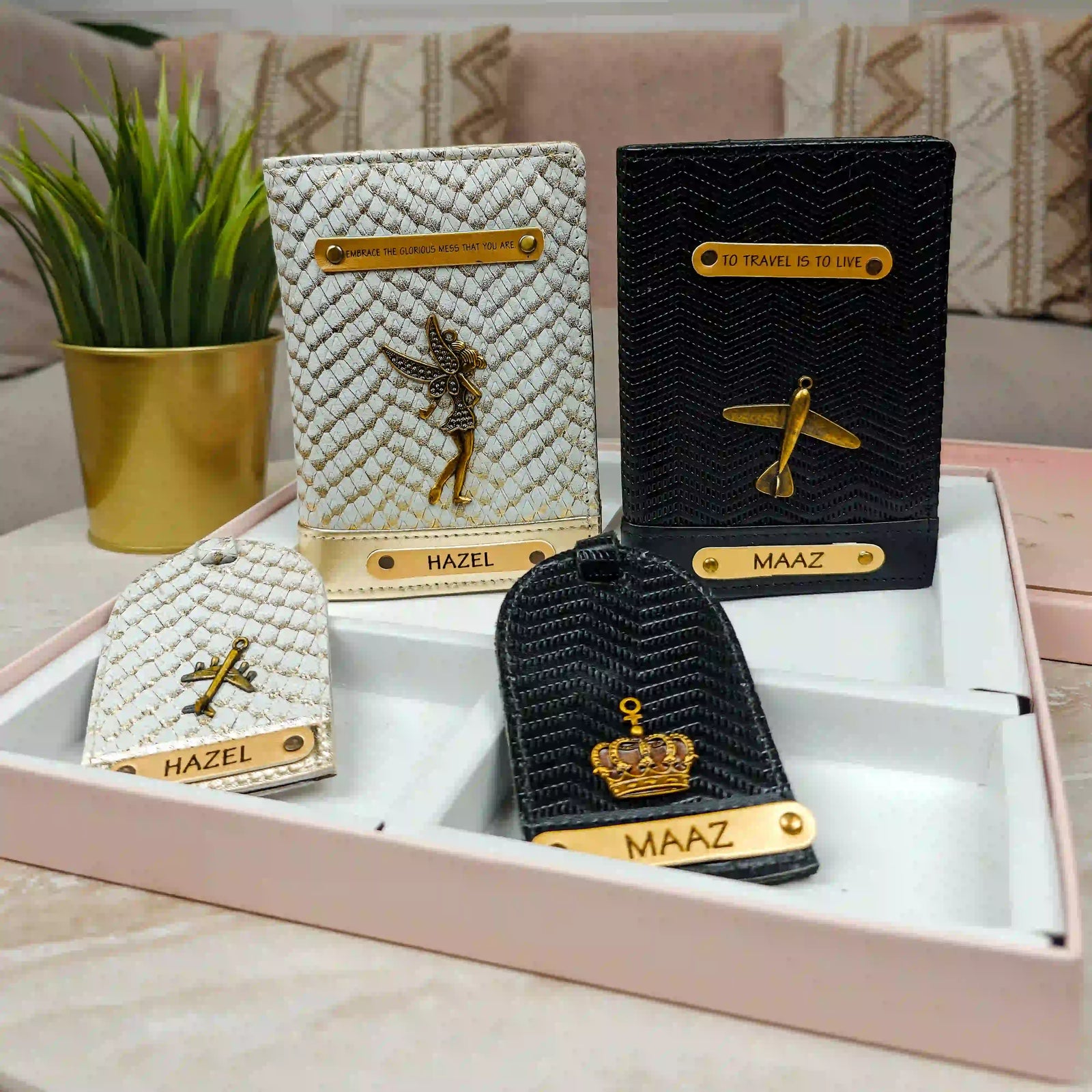 Personalised passport cover and personalized leather luggage tags displayed in a pink gift box on a marble surface, with a potted plant and sofa in the background. The tags feature textured cream and black leather, gold-tone hardware, and custom names 'HAZEL' and 'MAAZ' with gold initials.