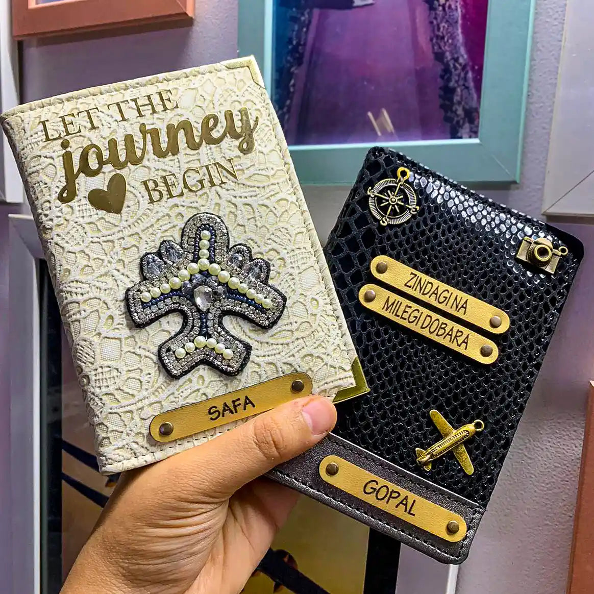 A hand holds two decorative passport covers in a real indoor setting with a framed picture in the background. One is white glided grace italian vegan leather passport cover with gold lettering reading 'LET THE journey BEGIN' & the other is black bollywood genuine leather passport cover with gold nameplates and a gold airplane charm - TPC Gifts