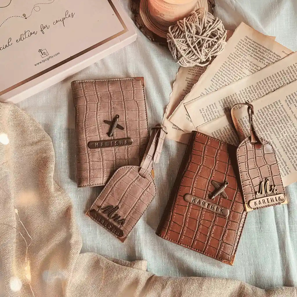 Two personalized leather luggage tags and matching passport holder set, one in light taupe and one in medium brown, styled on a light blue fabric with vintage papers, a candle, and fairy lights.