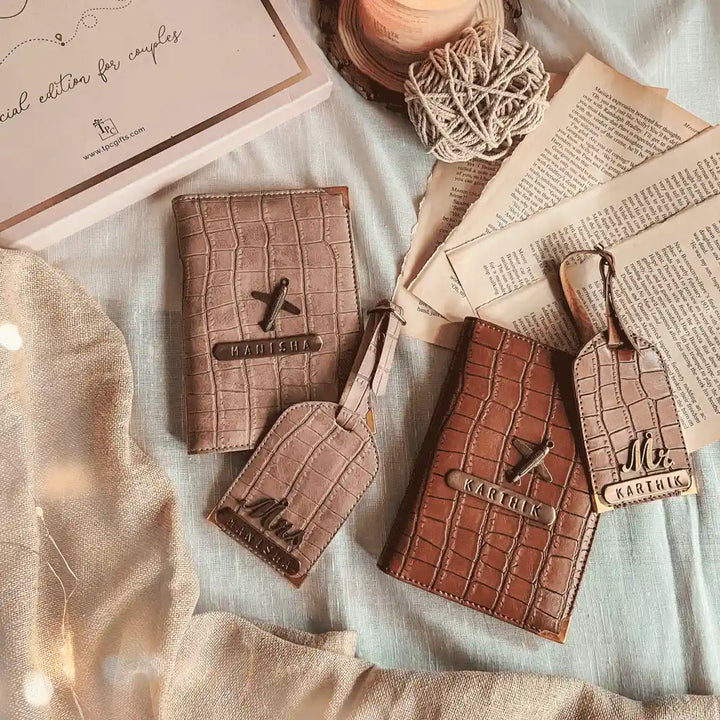 Two personalized leather luggage tags and matching passport holder set, one in light taupe and one in medium brown, styled on a light blue fabric with vintage papers, a candle, and fairy lights.