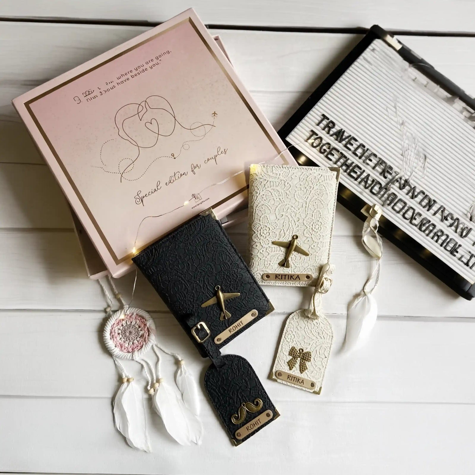 Two passport covers and two luggage tags with personalized engravings, one in black and one in a pastel color, displayed on a table with a pink gift box and a note reading 'Together Forever' in the background.