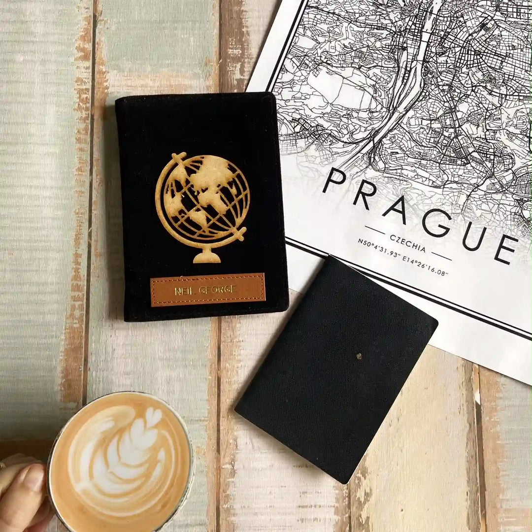 A black passport-style envelope with a gold globe emblem and a small brown leather label sits on a light wood surface next to a black notebook, a printed Prague map, and a cup of latte with latte art.
