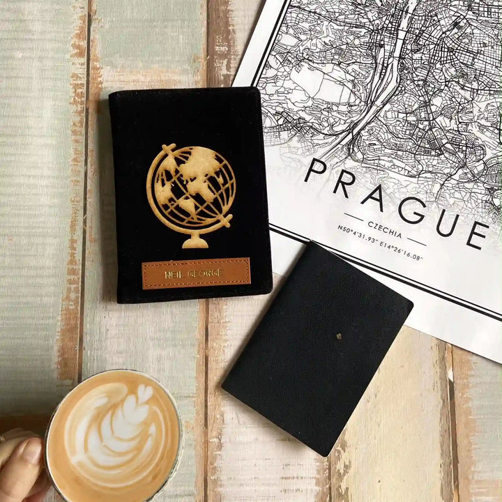 A black passport-style envelope with a gold globe emblem and a small brown leather label sits on a light wood surface next to a black notebook, a printed Prague map, and a cup of latte with latte art.