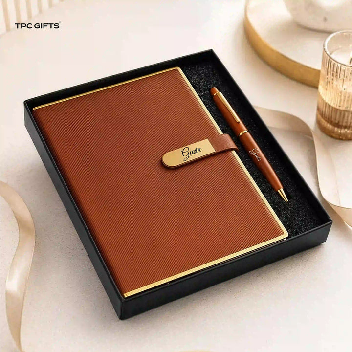 A brown textured notebook with a gold-accented strap and a matching brown pen presented in a black gift box. The scene is styled on a light surface with ribbon and a candle in the background.
