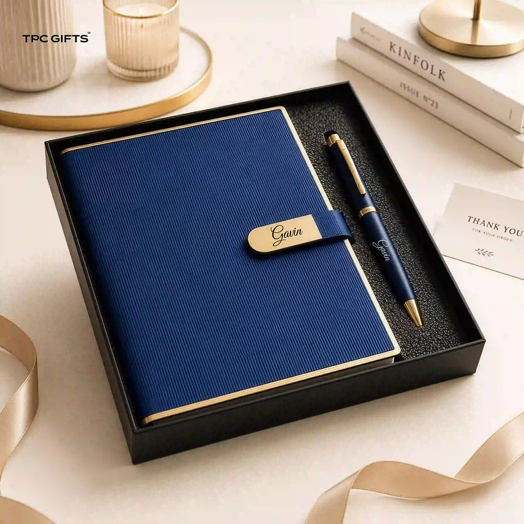 A navy blue textured notebook with a matching pen presented in a black gift box. The notebook has a gold trim and a gold nameplate reading &
