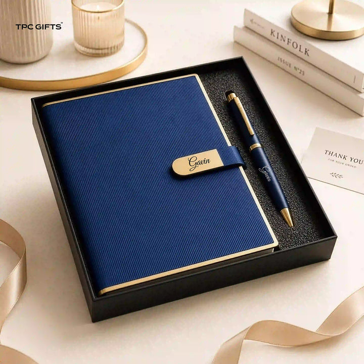 A navy blue textured notebook with a matching pen presented in a black gift box. The notebook has a gold trim and a gold nameplate reading &