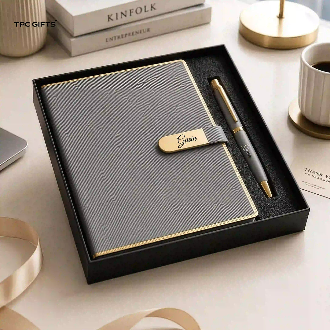 A gray textured notebook with a gold-embossed strap and pen set inside a black gift box, styled on a light beige desk with books, a coffee cup, and ribbon.