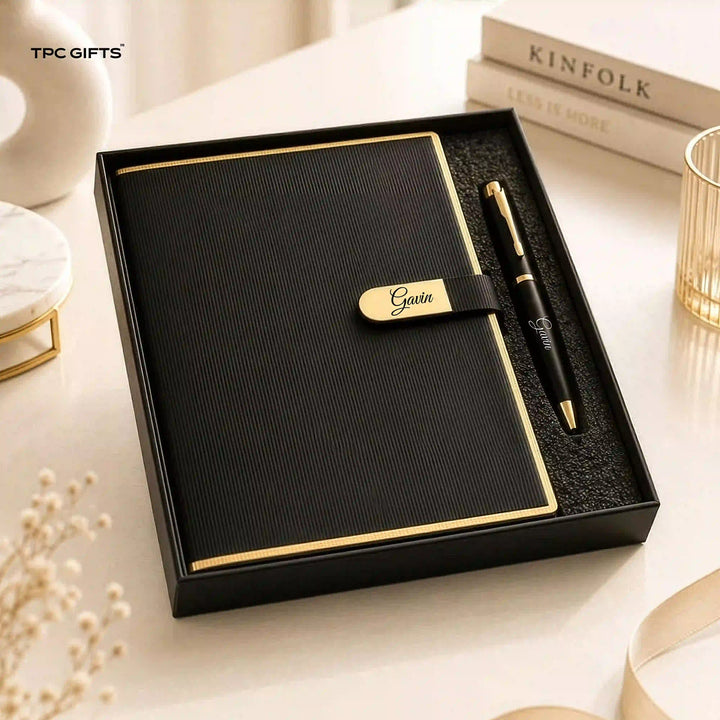 A black textured gift set box containing a black notebook with gold trim and a matching black pen with gold accents. The notebook has a black strap with the name &