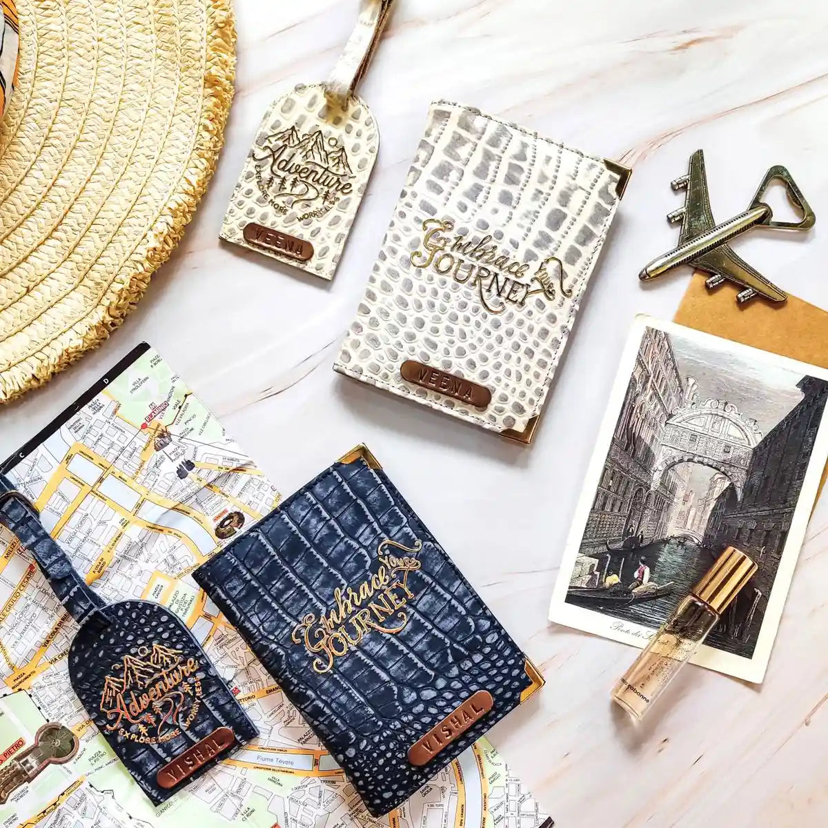 A styled flat-lay of two embossed genuine crocodile leather travel accessories on a light marble background, alongside a straw hat, vintage map, antique keys, a postcard, and a perfume bottle. The set includes a navy blue set and a cream/beige set, both with gold foil 'Eternal Adventure JOURNEY' lettering and brown leather brand tabs.