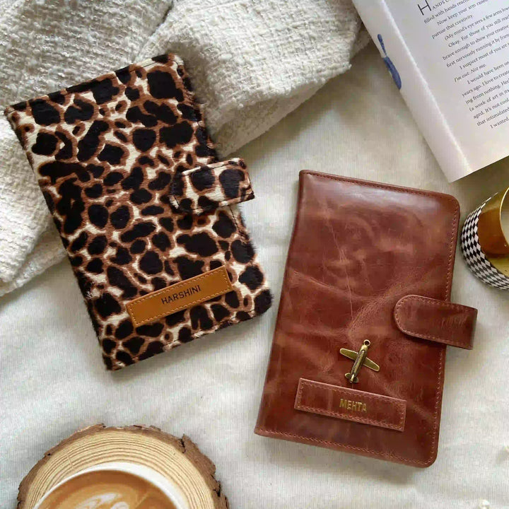 Two personalized planner covers styled on a textured white fabric background with lifestyle props. One cover features a brown and black leopard print pattern with a tan leather tab labeled &