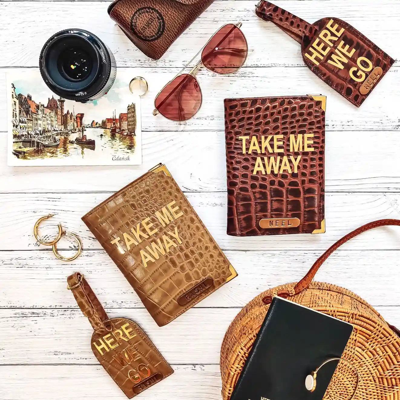 A styled flat-lay of crocodile-embossed leather accessories on a white wood background, including two wallets with 'TAKE ME AWAY' text, two bottle-shaped keychains with 'HERE WE GO' text, sunglasses, a camera lens, a postcard, rings, and a woven bag.