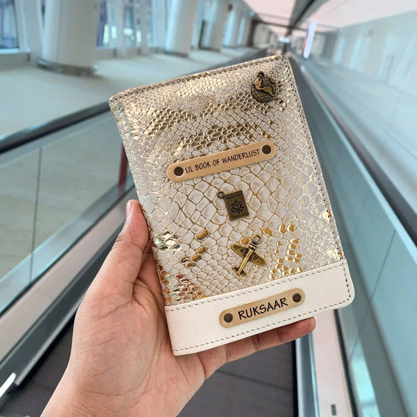 A hand holding a white personalized passport cover with gold details and a name tag that reads 'Book of Wanderlust'. - TPC Gifts