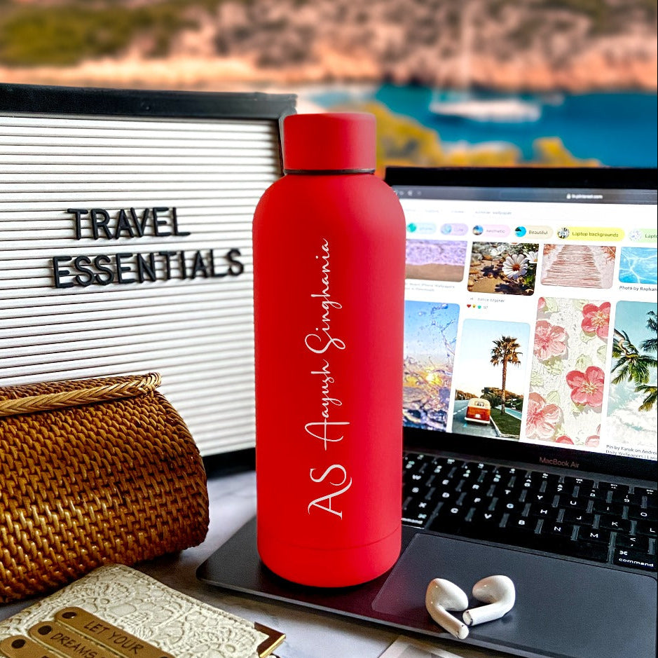A cherry red stainless steel water bottle with custom engraving, displayed on a desk alongside a laptop and a beach-themed travel essentials board by TPC Gifts