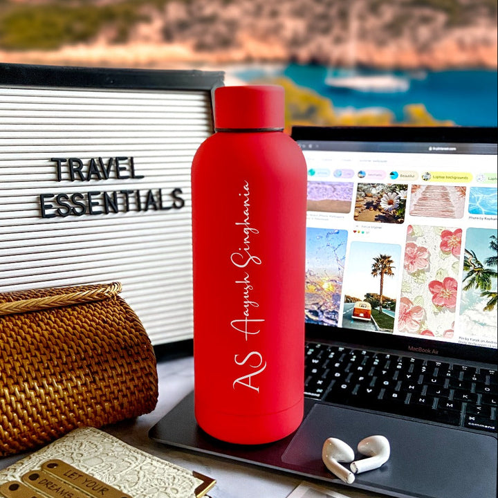 A cherry red stainless steel water bottle with custom engraving, displayed on a desk alongside a laptop and a beach-themed travel essentials board by TPC Gifts