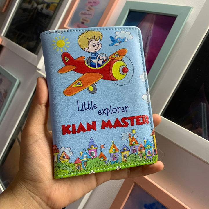 A hand holding a blue kids passport cover with an illustration of an airplane and the text &