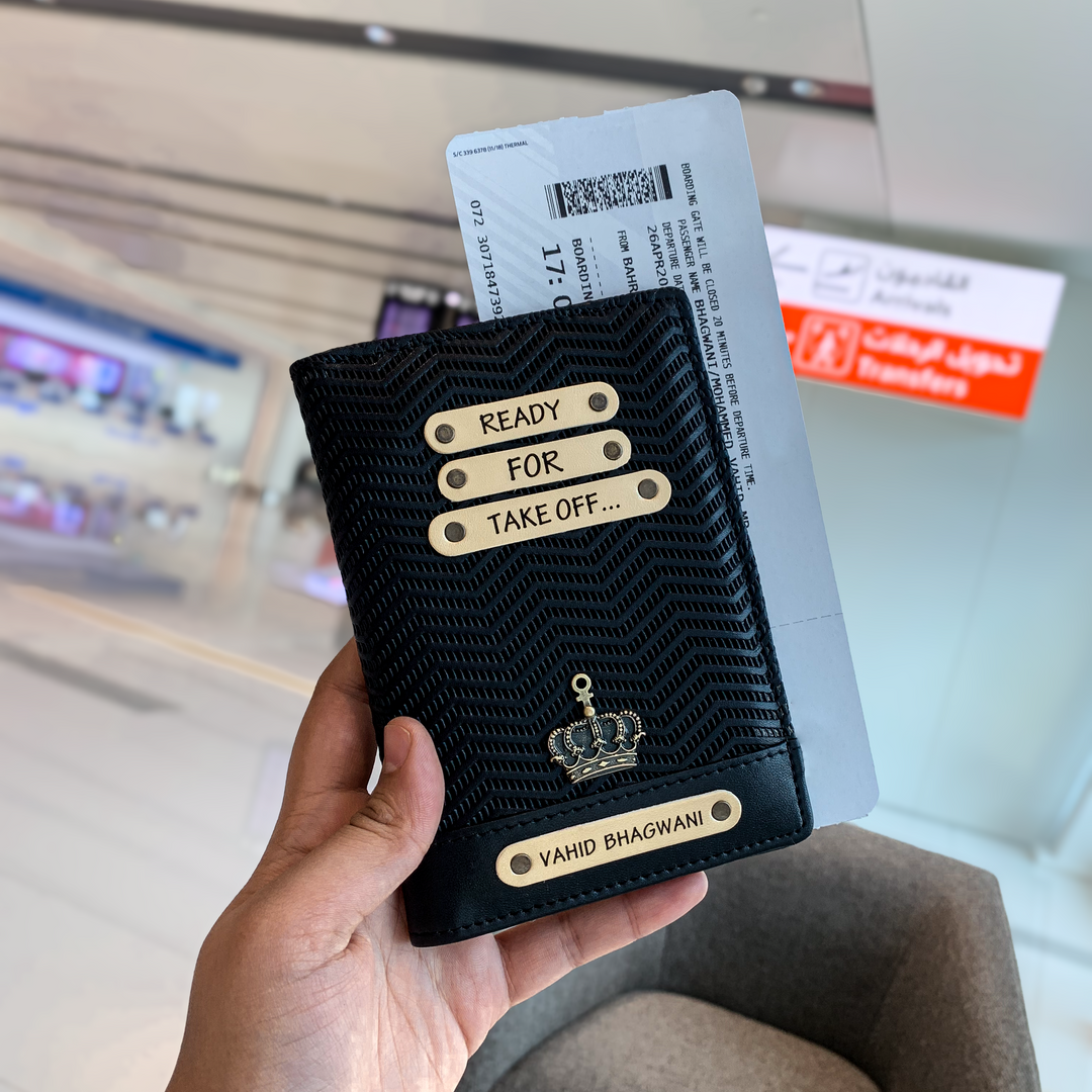 A hand holding a black personalized passport cover with text on it, displayed in an airport setting.